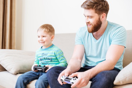 Happy cute son sitting on couch and playing computer games  with his father at home