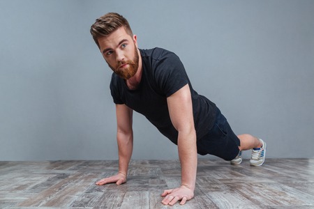 Serious bearded young sportsman doing plank core exercise over grey background