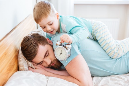 Cute little son holding alarm clock near sleeping father ear at homeの写真素材