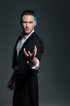 Attractive mysterious young magician holding something on palm over grey backgroundの写真素材