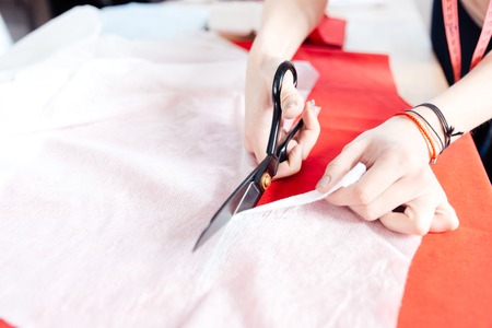 Hands of young woman seamstress with scissors cutting white fabric