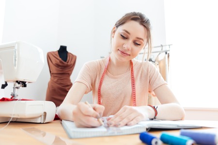 Smiling pretty young woman seamstress drawing pattents in notebook at work