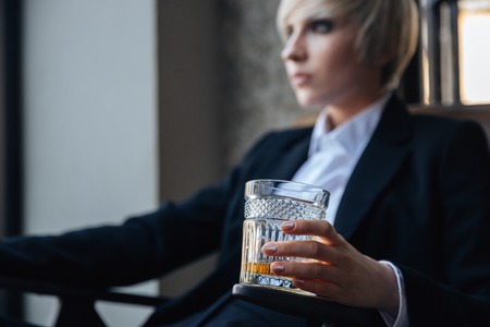 Stylish blonde girl sitting in chair in cafe with glass of whiskeyの写真素材
