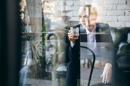 Stylish pretty blonde girl holding a glass of alcohol drink in a cafe, street reflection in windowの写真素材