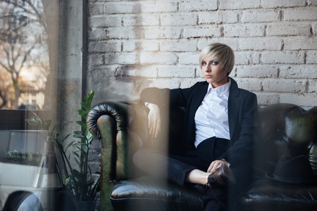 Blonde girl sitting on the couch and looking seriously in the cafe, street reflection in windowの写真素材