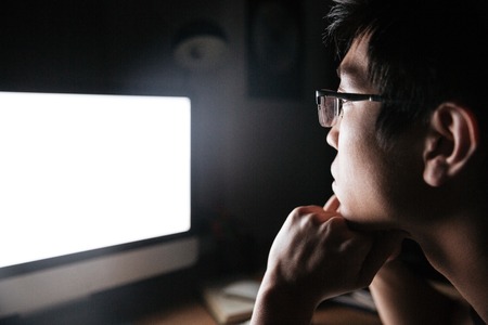 Concentrated asian young man in glasses looking at blank screen of computer in dark roomの写真素材