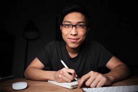 Happy attractive asian young man in glasses writing and smiling in dark roomの写真素材