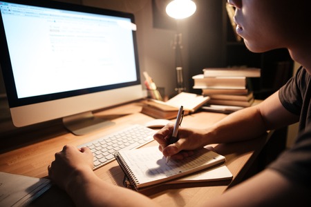 Closeup of concentrated young man writing in notepad and using computer in dark room at homeの写真素材