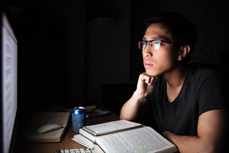 Serious asian young man sitting and studying with books and computer in dark room