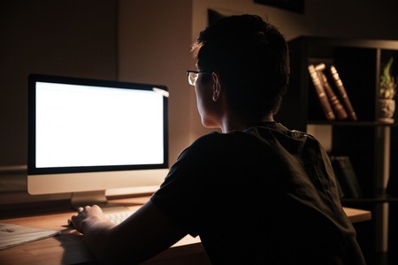 Back view of serious young man in glasses sitting and working with blank screen computer at home at...