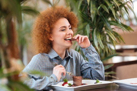 Happy beautiful redhead young woman sitting in cafe and eating chocolate dessertの写真素材