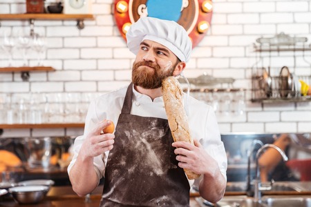 Pensive bearded baker holding eggs and bread and thinking on the kitchenの写真素材