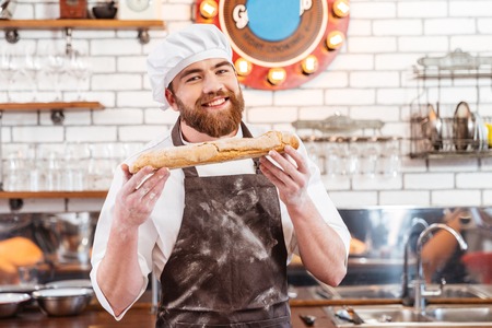 Cheerful attractive baker standing and showing loaf of bread on the kitchenの写真素材