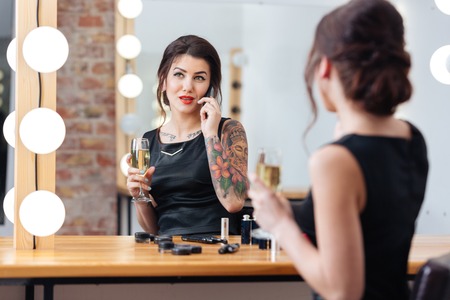 Charming young woman drinking champagne and talking on mobile phone in dressing roomの写真素材