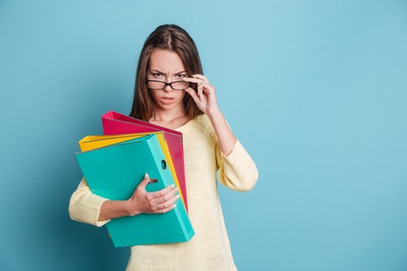 Seriously looking smart girl holding colorful binders isolated on the blue backgroundの写真素材