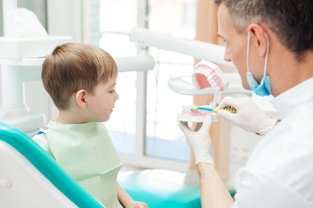 Dentist educating little boy about brushing teeth demonstrating on dental jaw model in clinicの写真素材