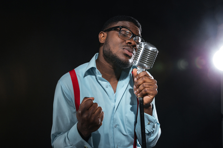 Handsome afro amerian man singing into vintage microphone over dark backgroundの写真素材