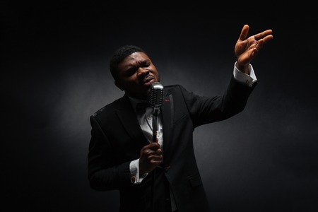 Handsome afro amerian man singing into vintage microphone over dark backgroundの写真素材