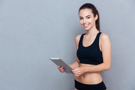 Beautiful young sportswoman using pc tablet and looking at camera isolated on grey backgroundの写真素材