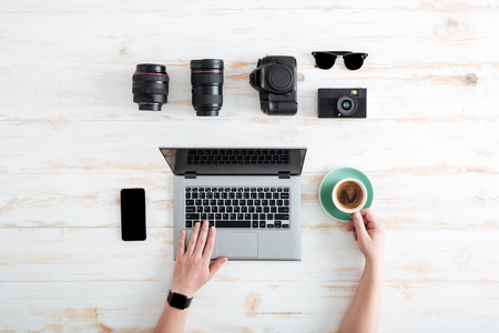 Hands of young man using laptop and drinking coffee on wooden table with blank screen mobile phone, sunglasses, lenses and photo cameraの写真素材