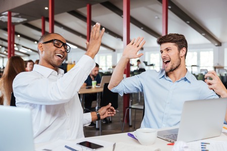 Two cheerful excited young men giving high five and working in office togetherの写真素材