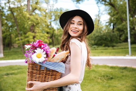 Smiling attractive young woman in hat holding basket with flowers, drinks and foodの写真素材