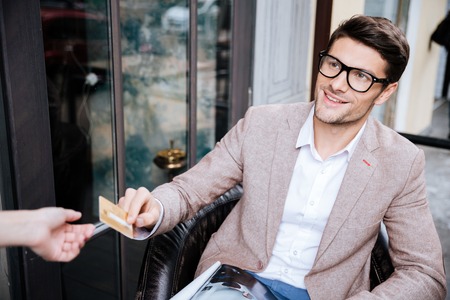 Happy young man in glasses sitting in outdoor cafe and paying by credit cardの写真素材