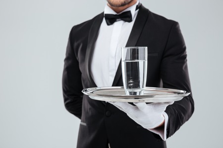 Closeup of butler in tuxedo and gloves holding silver tray with glass of waterの写真素材
