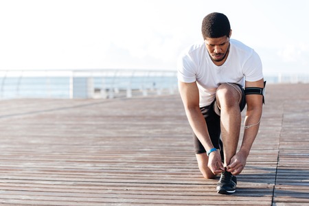 Serious african american young sportsman laces his sneakers on wooden terraceの写真素材