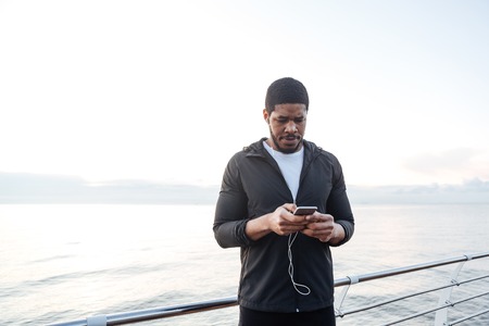 Serious african young sportsman listening to music from cell phone on pier