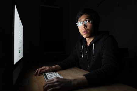 Amazed asian young man in glasses working with computer in dark office