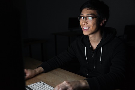 Cheerful asian young man in glasses using laptop in dark room