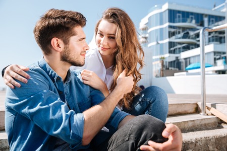 Portrait of a smiling young couple looking at each other and sitting at the seasideの写真素材