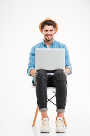 Smiling young man in hat sitting on chair and using laptop over white backgroundの写真素材