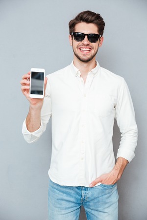 Happy handsome young man in sunglasses holding blank screen cell phone over grey backgroundの写真素材