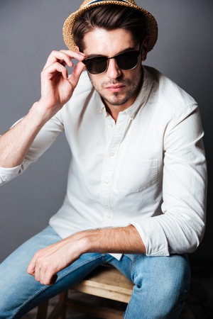 Fashion portrait of serious young man in white shirt, jeans, sunglasses and hat over grey backgroundの写真素材