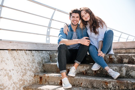 Portrait of a smiling happy young couple looking at camera and sitting at the seasideの写真素材