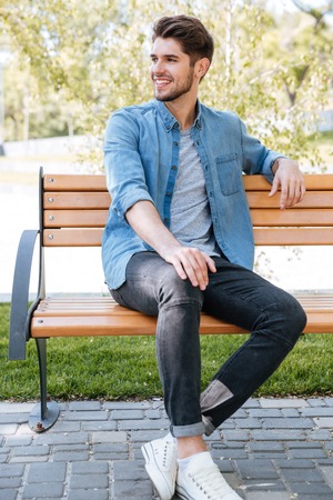 Handsome young man sitting on a bench smiling in the parkの写真素材