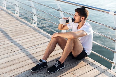Attractive young male athlete sitting on pier and drinking waterの写真素材