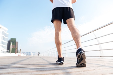 Back view of legs of young man in black shorts and sneakers running on pier
