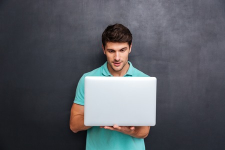 Concentrated handsome young man standing and using laptop over chalkboard background