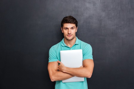 Serious confident young man standing and holding laptop over blackboard backgroundの写真素材