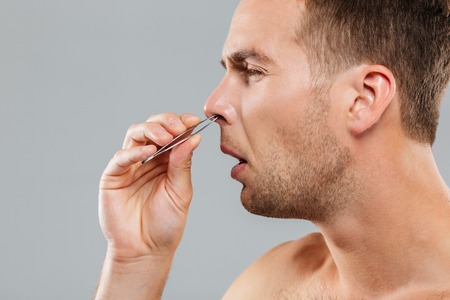 Side view of a man removing nose hair with tweezers isolated on the gray backgroundの写真素材