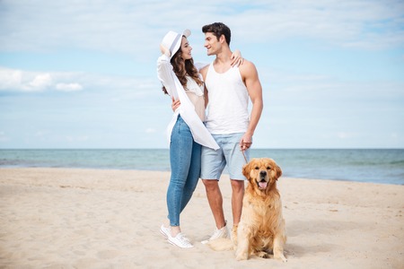 Happy young couple hugging and smiling while walking with dog on the beachの写真素材