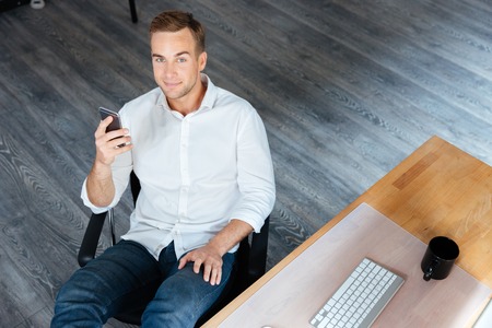 Top view of smiling relaxed young businessman sitting and using smartphone in office