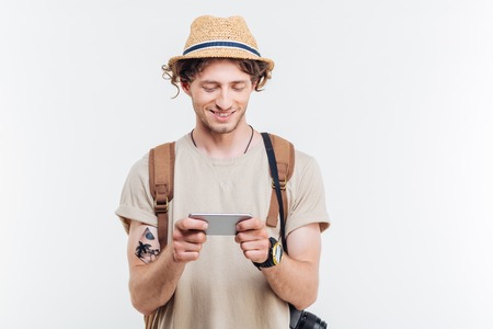 Portrait of a smiling young man with backpack and camera using smart phone isolated on a white backgroundの写真素材