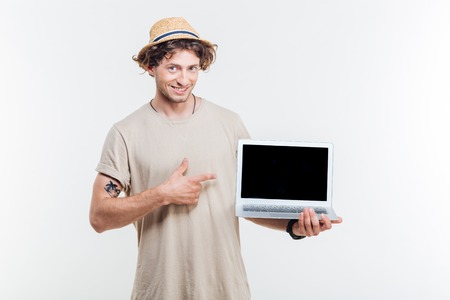 Smiling attractive young man holding blank screen laptop and pointing at it over white backgroundの写真素材