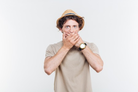Handsome young man in hat coveres his mouth by hands over white backgroundの写真素材