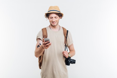 Portrait of a smiling young man with backpack and camera using smart phone isolated on a white backgroundの写真素材