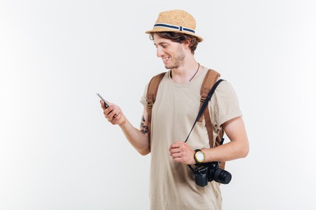 Portrait of a smiling young man with backpack and camera using smart phone isolated on a white backgroundの写真素材
